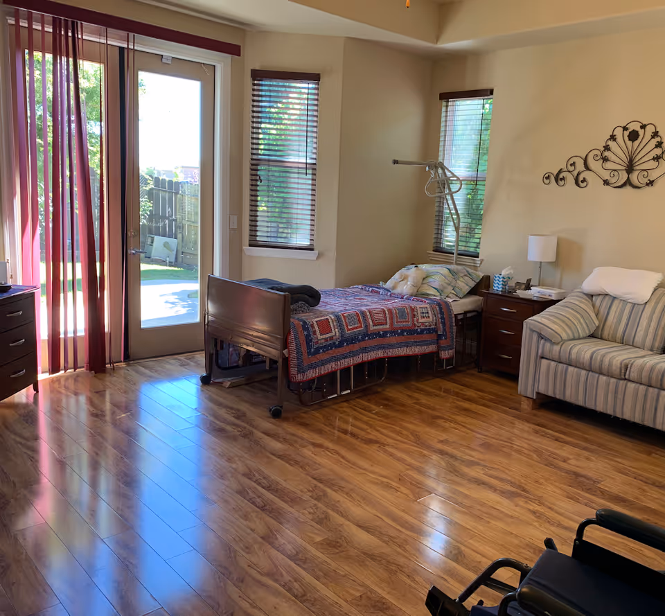 A bright room with wooden flooring featuring a hospital-style bed covered with a colorful quilt, a striped armchair, a wooden nightstand with a lamp and tissues, and a wheelchair in the foreground. There are two windows with blinds and a glass door with sheer curtains leading outside to a garden area.