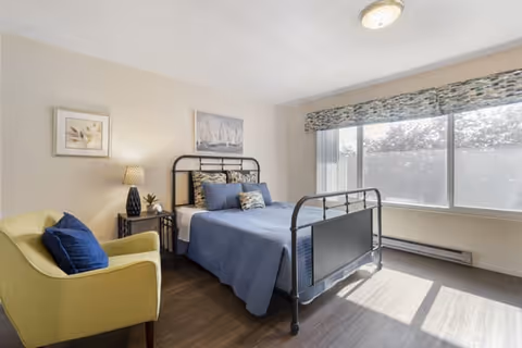 A bright bedroom with a metal-framed bed dressed in blue bedding and multiple pillows. There is a yellow armchair with a blue cushion to the left, a small side table with a lamp and plant, and two framed pictures on the wall. Large windows with patterned valances allow natural light to fill the room.