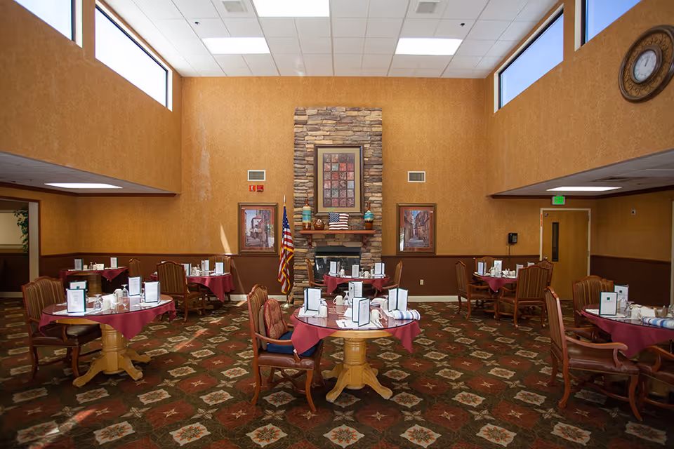Dining room with several round tables covered with burgundy tablecloths, each set with menus, napkins, and glassware. The room has a high ceiling with two large windows near the top, a stone fireplace in the center of the far wall, and an American flag beside it. The walls are painted in warm tones, and there is a patterned carpet on the floor. Framed artwork and a clock are visible on the walls.