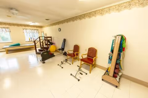 A spacious rehabilitation room with exercise equipment, therapy balls, weights, two chairs, and a small set of stairs along a cream-colored wall.