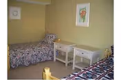 A small bedroom with two single beds placed against opposite walls. Each bed has a patterned bedspread, one floral and the other geometric. Between the beds are two white nightstands, one with a lamp on it. The walls are painted light yellow, and a framed floral picture hangs above one of the nightstands.