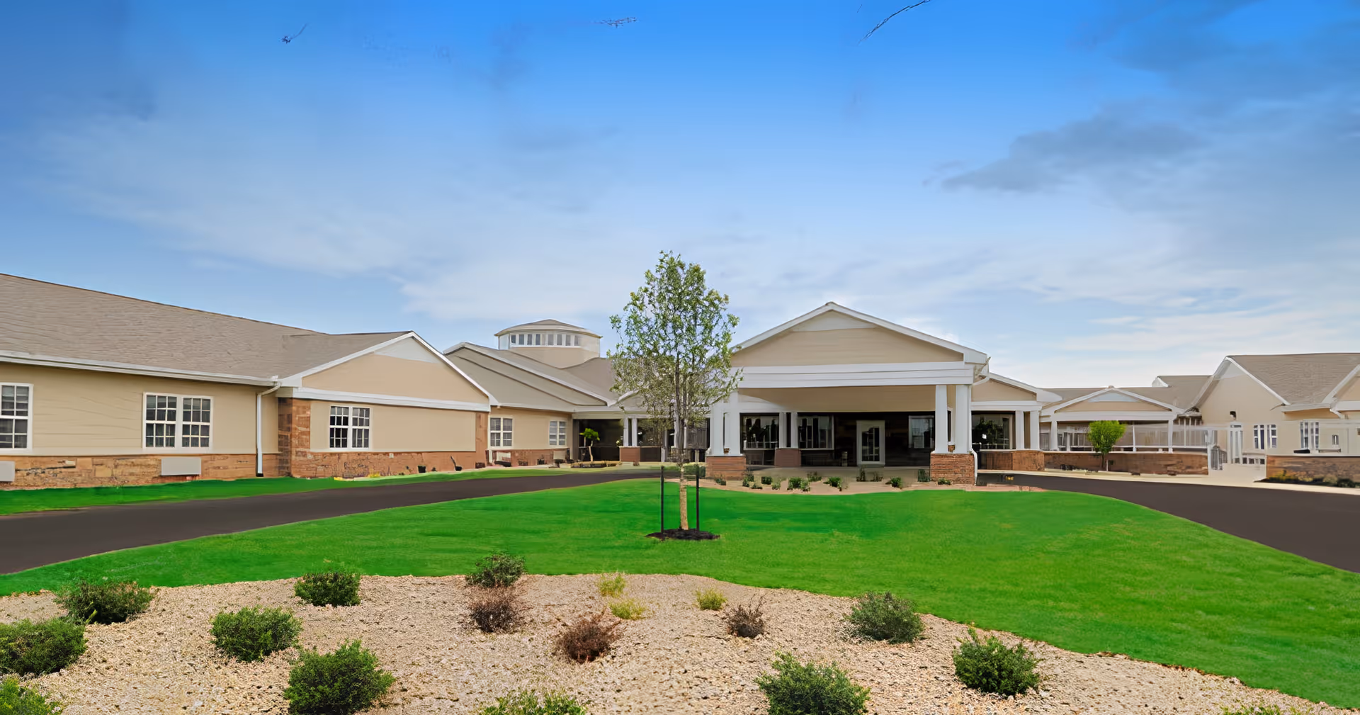Front exterior view of Cypress Pointe Health Campus showing a large building with beige walls, multiple windows, a covered entrance supported by white columns, a well-maintained green lawn, small shrubs, and a tree in the center under a partly cloudy blue sky.