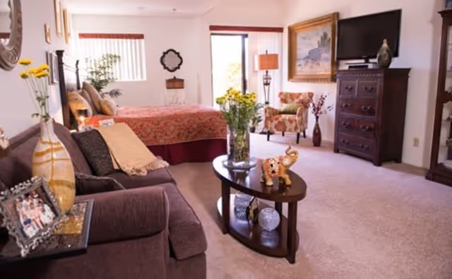 A cozy and well-decorated retirement facility room featuring a bed with a red patterned bedspread, a brown sofa with cushions and a throw blanket, a wooden coffee table with decorative items including a vase of yellow flowers and a small elephant figurine, an armchair with floral upholstery, a wooden dresser with a flat-screen TV on top, and framed artwork on the walls. The room has carpeted flooring and a window letting in natural light.