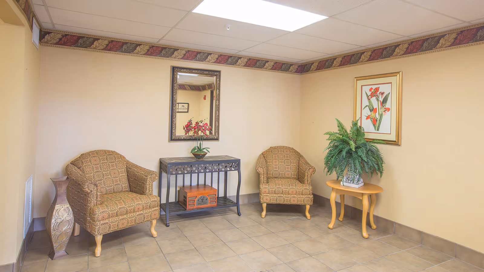 Small interior waiting area with two patterned armchairs, a console table and mirror, a side table with a plant, and framed floral artwork on the wall.