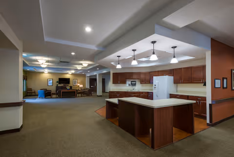 Spacious interior area featuring a modern kitchen with a large island, wooden cabinets, white refrigerator, and pendant lighting. In the background, there is a living room area with seating, a television, and framed artwork on the walls.