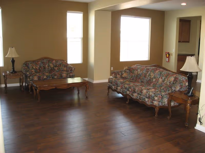 A living room with two floral patterned sofas facing each other, a wooden coffee table between them, and two wooden side tables each with a lamp. The room has wooden flooring, beige walls, and two windows letting in natural light.