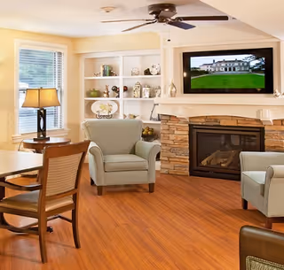 A cozy living room area with wooden flooring, two light green armchairs, a wooden dining table with chairs, a stone fireplace with a TV mounted above it, and built-in white shelves displaying decorative items. A window with blinds and a table lamp are also visible.