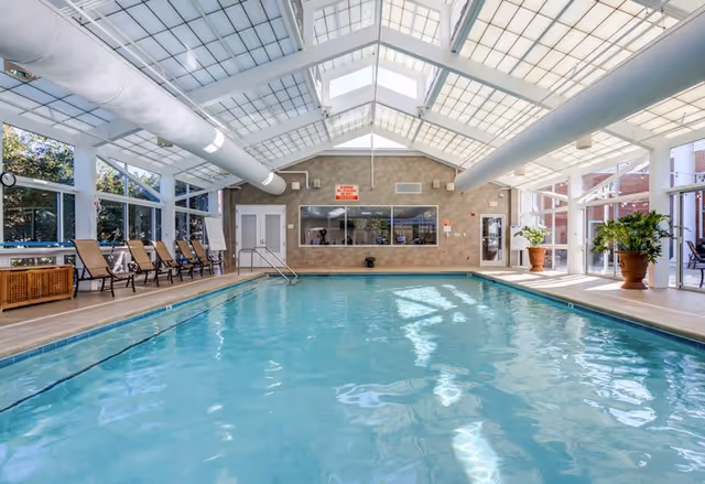 Indoor swimming pool with clear blue water, surrounded by lounge chairs and large windows letting in natural light. The ceiling is made of glass panels with white framing, and there are potted plants on the pool deck.