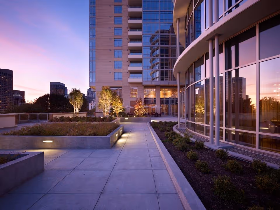 Outdoor terrace area of a modern high-rise building at dusk with illuminated trees, landscaped planters, and large glass windows reflecting the sunset sky.