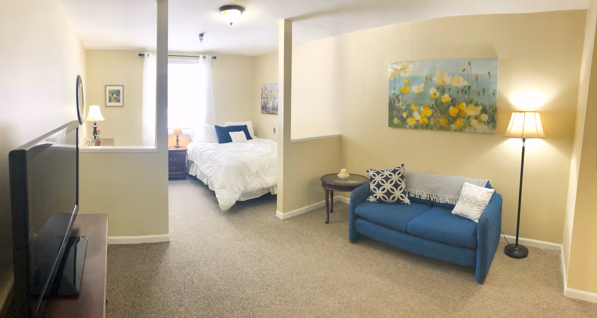 A cozy assisted living room and bedroom area with beige walls and carpet. The living room features a blue loveseat with patterned pillows, a small round wooden side table, a floor lamp, and a floral painting on the wall. The bedroom area has a bed with white bedding, a nightstand with a lamp, and a window with white curtains letting in natural light. A flat-screen TV is visible on a stand in the foreground.