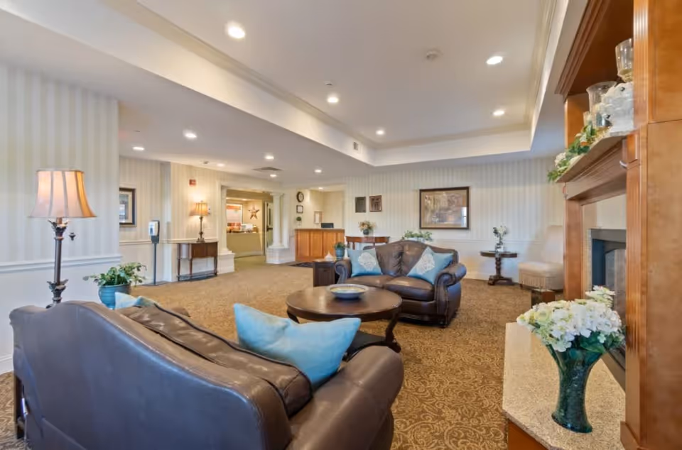 A spacious and well-lit living room area with two dark leather sofas adorned with blue and white pillows, a round wooden coffee table, a fireplace with decorative items and flowers, a floor lamp, and a reception desk in the background. The room has patterned carpet flooring and striped wallpaper.