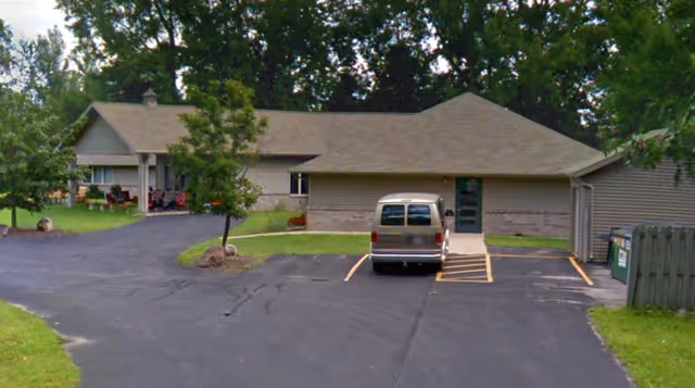 Single-story building with beige siding and a brown shingled roof, surrounded by green grass and trees. A gray van is parked in a marked parking space in front of the building. There is a paved driveway and parking area leading to the entrance, which has a green door. A small porch area with chairs is visible on the left side of the building.