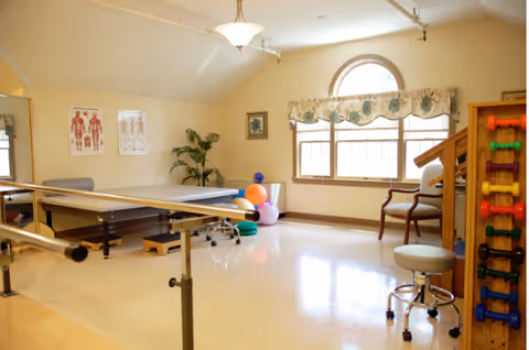 A rehabilitation room with parallel bars for walking exercises, colorful exercise balls, small dumbbells on a rack, anatomical charts on the wall, a chair near a large window with a floral valance, and a potted plant in the corner.