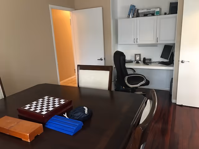 A room with a dark wooden table and chairs, a chessboard on the table, and a small office area with a black office chair, white cabinets, and a computer in the background. The room has wooden flooring and an open door leading to a hallway with tiled flooring.