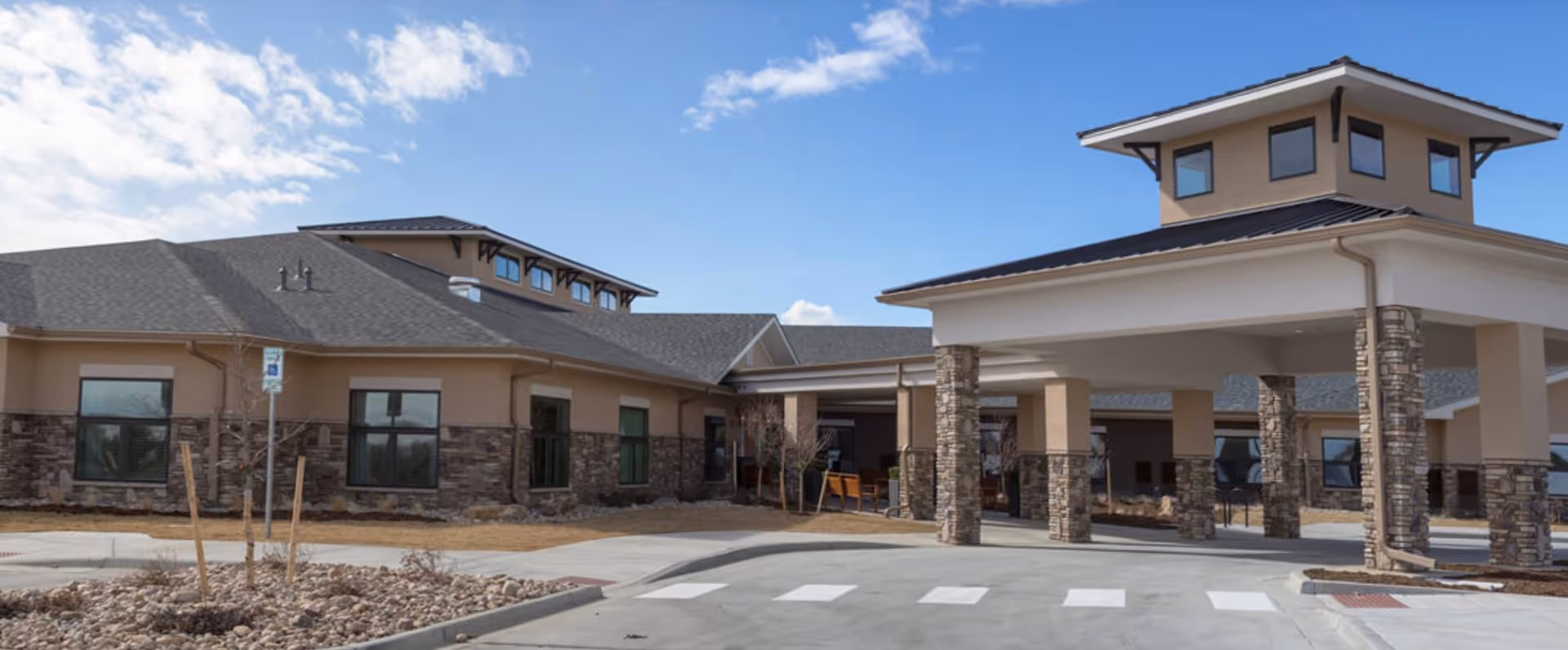 Exterior view of a single-story senior living facility building with stone and beige siding, a covered entrance supported by stone pillars, and a clear sky with some clouds.