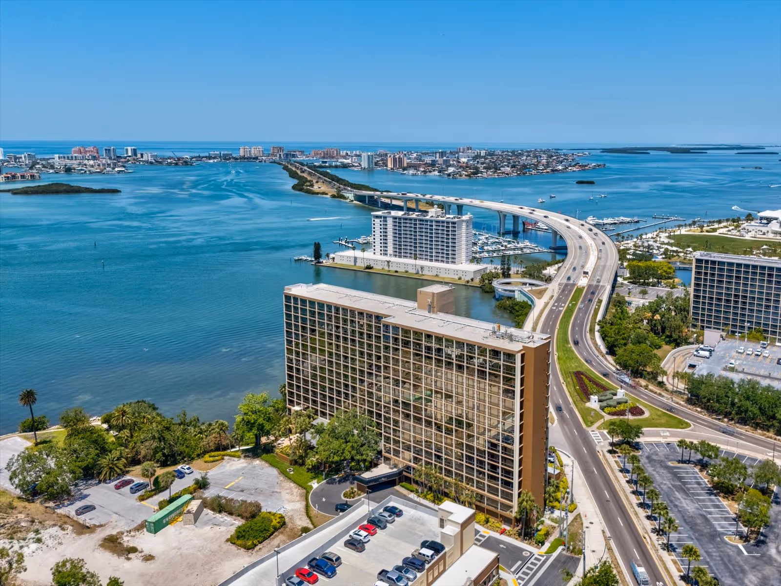 Aerial view of a waterfront area featuring a large multi-story building near a curved bridge over a body of water, with several cars parked in nearby parking lots and greenery surrounding the area under a clear blue sky.