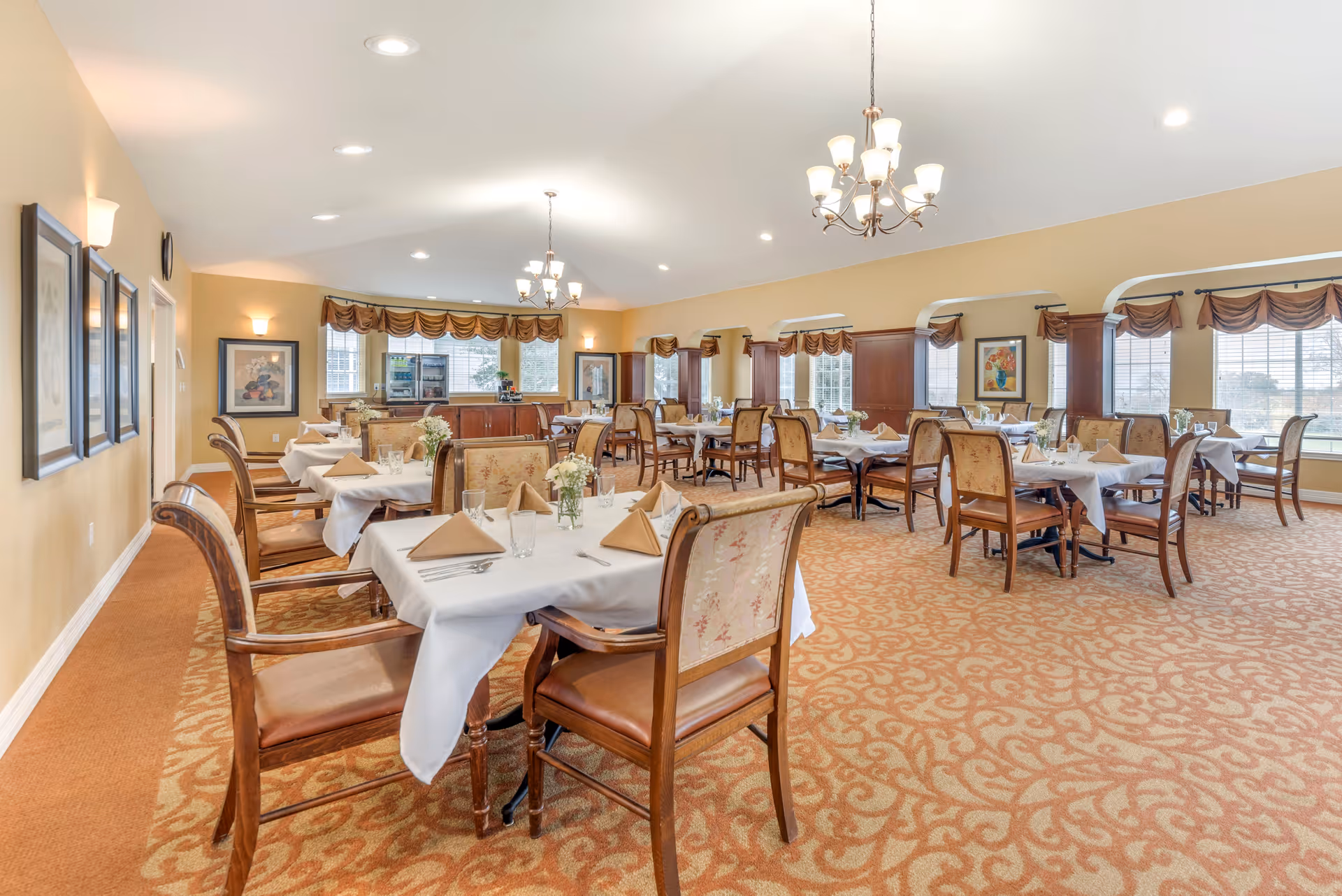 A spacious dining room with multiple tables covered in white tablecloths, each set with beige napkins, glassware, and silverware. The room features patterned carpet, wooden chairs with cushioned seats and backs, large windows with brown valances, chandeliers, and framed artwork on the walls.