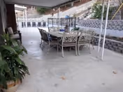 Covered outdoor patio area with a long dining table and multiple chairs arranged around it. The patio has a concrete floor and is adjacent to a retaining wall with plants and a hillside in the background.