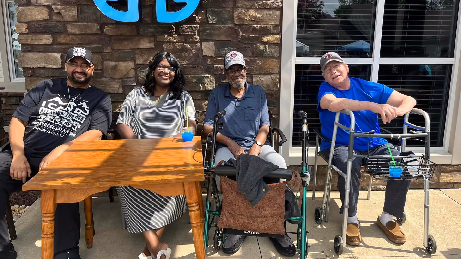 Four people sitting outside at a wooden table in front of a stone wall with a blue logo. Two men and one woman are seated on chairs, and one man is seated on a walker. They are smiling and appear to be enjoying a sunny day.