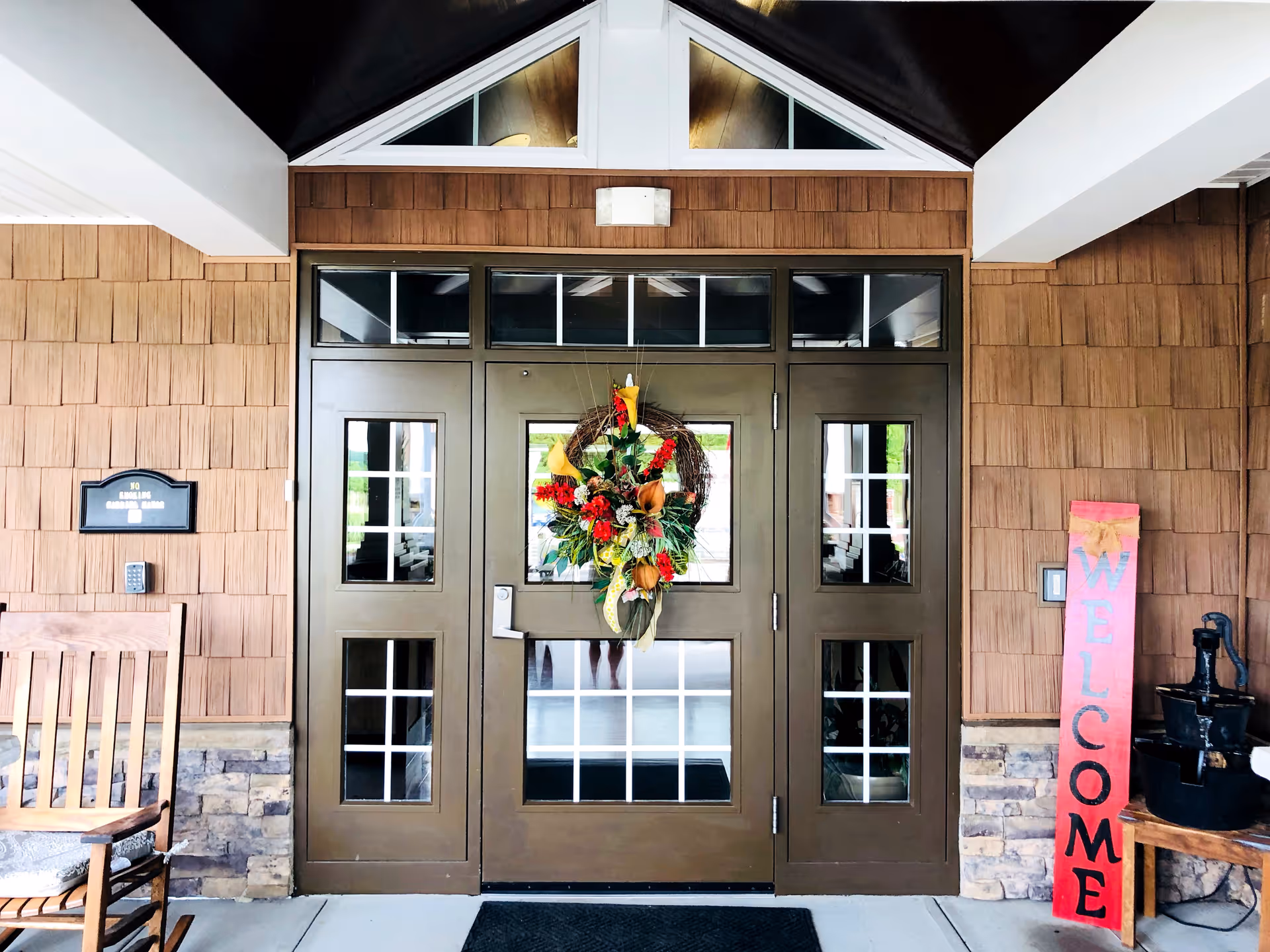 Entrance door of a building with a decorative wreath hanging on the door. The door is brown with multiple small windows. To the left, there is a wooden rocking chair and a plaque on the wall that reads 'Gabriel Manor'. To the right, there is a tall red wooden sign with the word 'WELCOME' written vertically and a small black water pump on a wooden table. The exterior wall is covered with brown shingles and stone accents at the bottom.