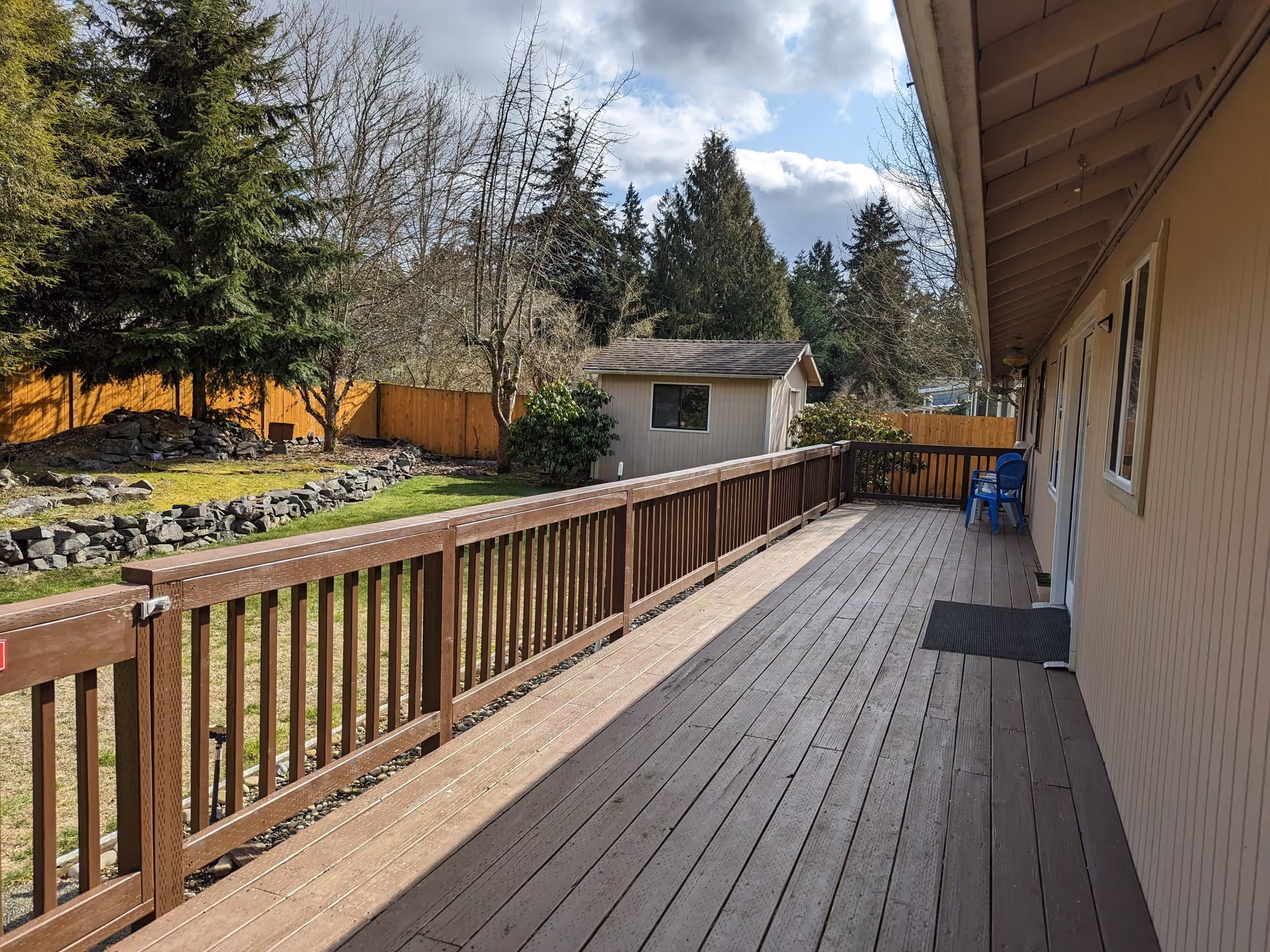 A wooden deck attached to a beige building with a brown railing. There are two blue chairs at the far end of the deck. Beyond the deck is a fenced yard with green grass, trees, and a small shed. The sky is partly cloudy.