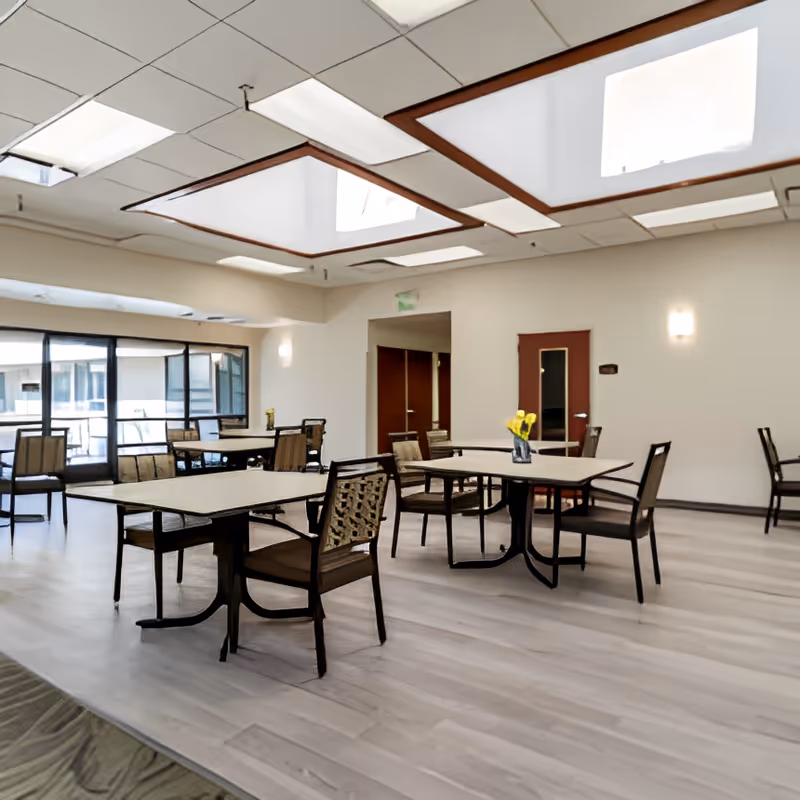 Bright communal dining room with tables and chairs under skylights and a vase of yellow flowers on a table.