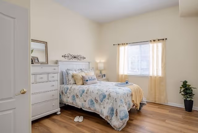 A bright bedroom with a bed covered in a floral comforter and multiple pillows, including one with the word 'believe'. There is a white dresser with a mirror and framed photos on top, a nightstand with a lamp and picture frame, a window with sheer curtains letting in natural light, wooden flooring, a pair of slippers on the floor, and a potted plant in the corner.