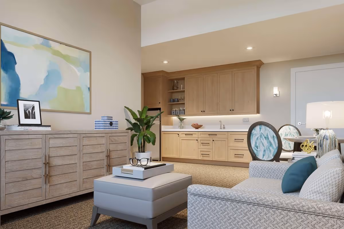 Bright living room with a sofa, ottoman, sideboard, artwork, potted plant and a kitchenette with wooden cabinets.