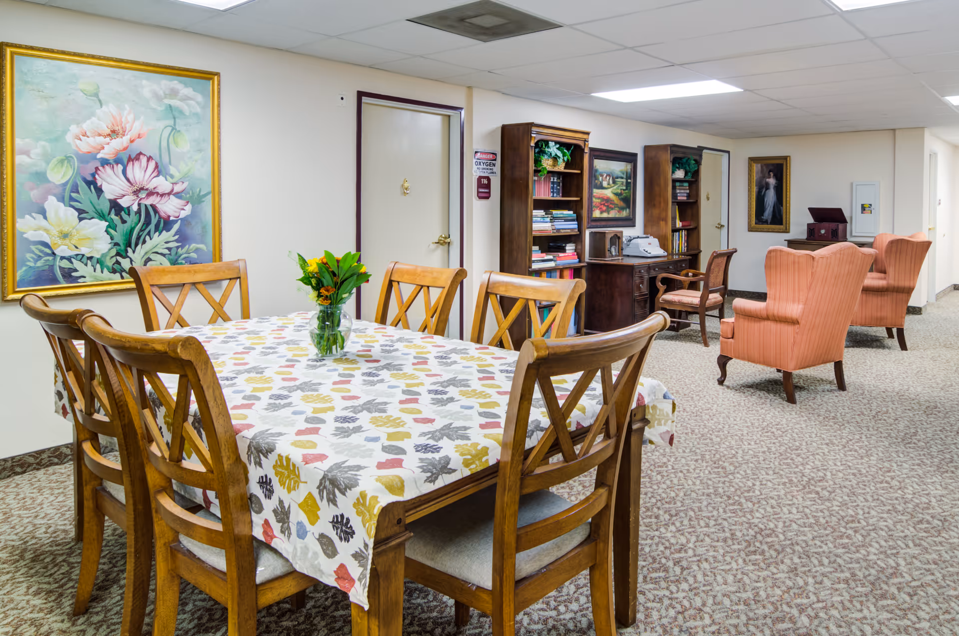 A cozy senior living common area featuring a wooden dining table with six chairs and a floral tablecloth, a vase with flowers on the table, two wooden bookshelves filled with books, a wooden desk with a typewriter, two upholstered armchairs, and framed paintings on the walls.