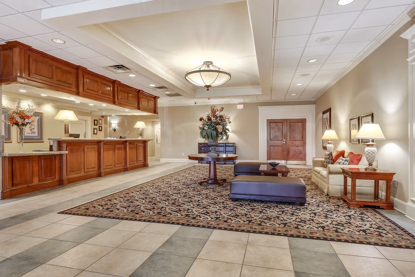 A spacious senior living facility lobby with a large patterned rug, a round wooden table with a floral arrangement in the center, a beige sofa with red and patterned cushions, two matching side tables with lamps, and a wooden reception desk with lamps and floral decor. The walls are light-colored with framed artwork, and the ceiling has recessed lighting and a central chandelier.