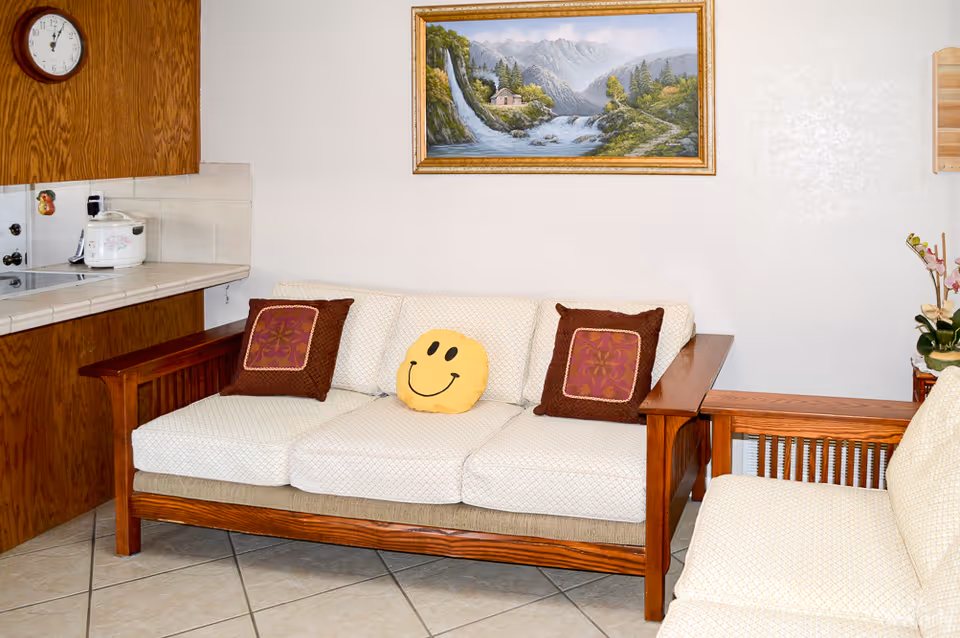 A cozy living room area with a wooden-framed beige sofa and matching armchair. The sofa has two brown decorative pillows and a yellow smiley face pillow. Above the sofa, there is a framed painting of a scenic mountain landscape with a waterfall and a small house. To the left, part of a kitchen counter with a rice cooker and a wall clock is visible. The floor is tiled and the walls are light-colored.
