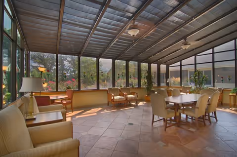 A bright sunroom with large windows and a glass ceiling allowing natural light to fill the space. The room contains several cushioned armchairs, a table with chairs, potted plants, and a tiled floor. Outside the windows, greenery and flowers are visible.