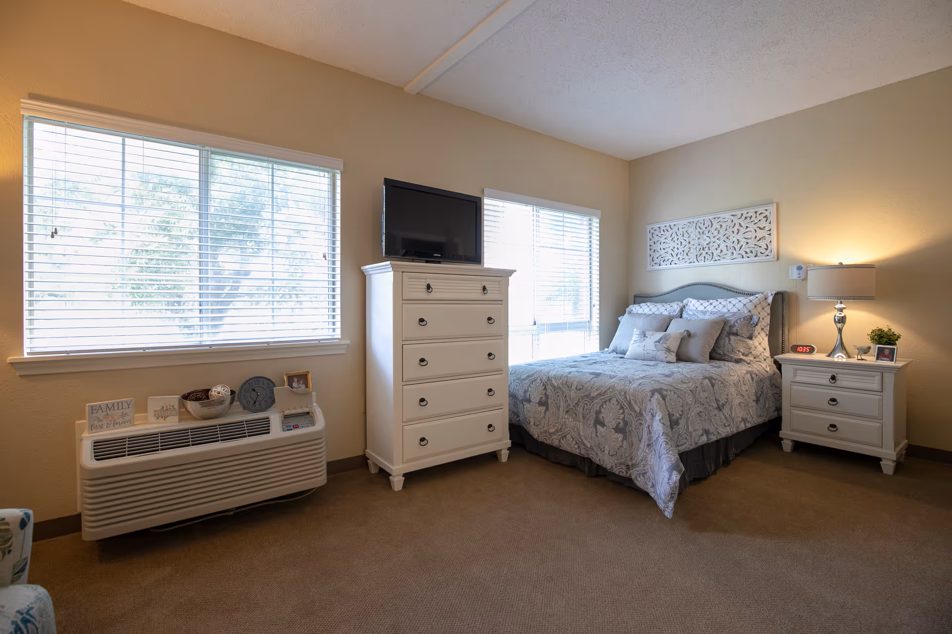 A cozy bedroom with a bed covered in patterned bedding and multiple pillows. Next to the bed is a white nightstand with a lamp, a digital clock, a small plant, and a framed photo. Across from the bed is a white chest of drawers with a flat-screen TV on top. The room has beige walls, carpeted floor, and two large windows with blinds letting in natural light. A decorative wall piece hangs above the bed.