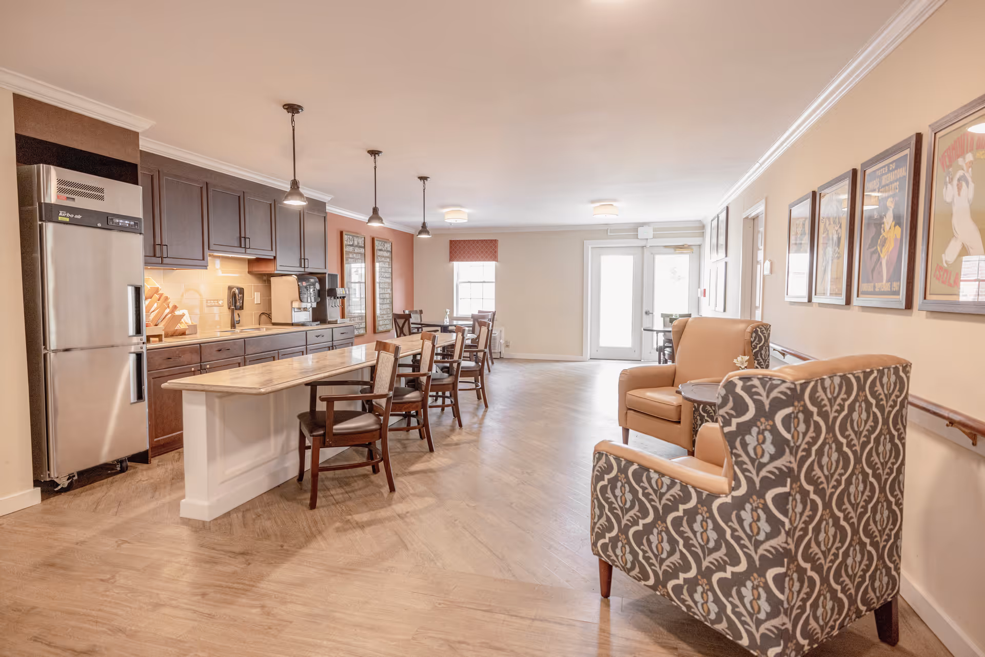 A bright and spacious common area in a senior living facility featuring a kitchen with dark wood cabinets, a stainless steel refrigerator, and a marble countertop island with chairs. The room also includes a seating area with patterned armchairs and a small round table, framed artwork on the walls, and large windows letting in natural light.