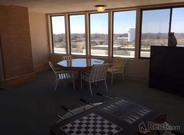 A senior living facility common room with large windows showing an outdoor view. The room contains a round wooden table with four metal chairs around it, a foosball table with a chessboard on top, and a dark cabinet with a decorative owl on it. The floor is carpeted and there is a brick column on the left side.