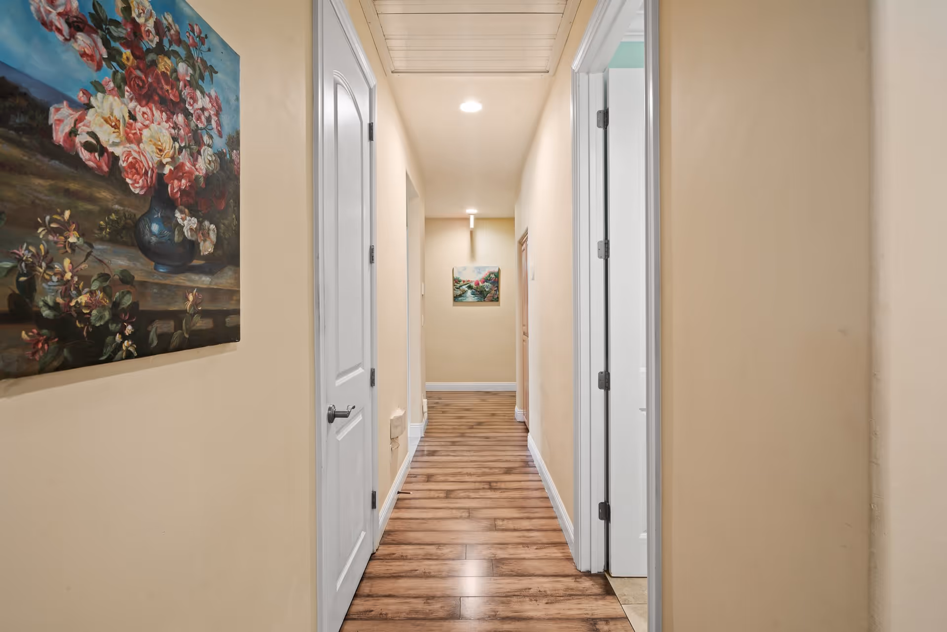 A narrow hallway with wooden flooring and beige walls, featuring white doors on both sides and floral paintings hanging on the walls.