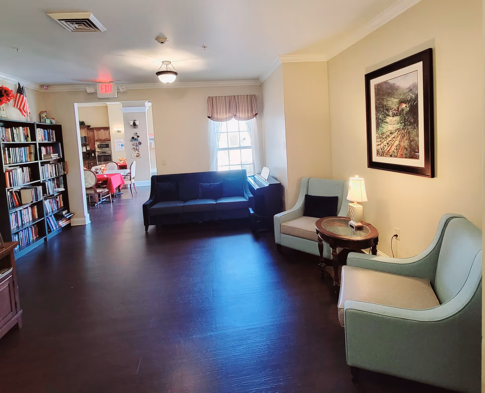 A cozy senior living common area with a dark blue sofa, two light blue armchairs, a round wooden side table with a lamp, a framed landscape painting on the wall, and a bookshelf filled with books. In the background, a dining area with a table covered in a red tablecloth and chairs is visible through an open doorway.
