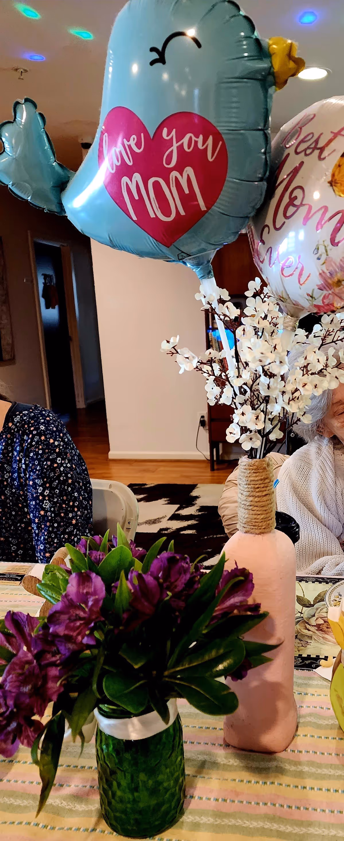 A close-up of a table decorated with two vases of flowers and two balloons. One balloon is blue with a pink heart that says 'love you MOM,' and the other is white with floral designs and the words 'Best Mom Ever.' Part of an elderly person wearing a white sweater is visible on the right side, and another person in a dark floral shirt is partially visible on the left. The setting appears to be a cozy indoor living space with wooden floors and a patterned rug.
