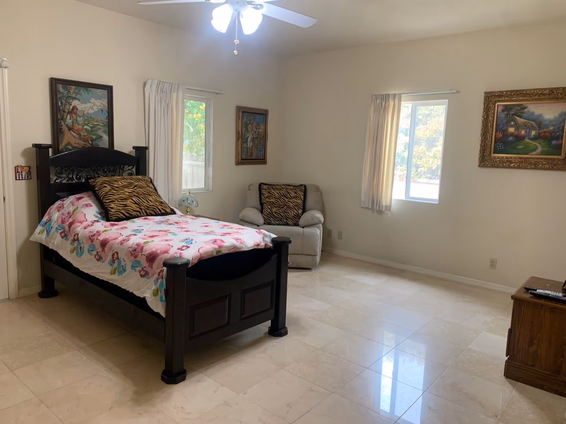A bedroom with a single dark wooden bed featuring a floral bedspread and a tiger-striped pillow. There is a beige armchair with a matching tiger-striped pillow near two windows with white curtains. The room has light-colored tiled flooring, three framed paintings on the walls, a ceiling fan with lights, and a wooden cabinet with a remote control on top.