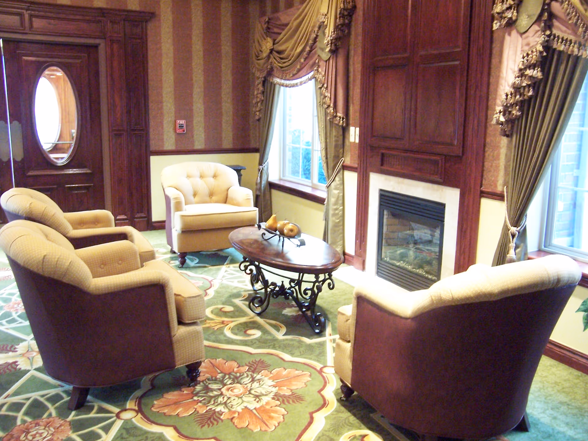 A cozy sitting area with four upholstered armchairs arranged around an oval wooden coffee table with decorative fruit. The room features a fireplace with a wooden mantel, large windows with ornate drapes, patterned carpet, and wood-paneled walls.