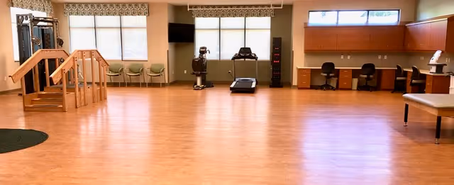 A spacious rehabilitation or physical therapy room with wooden flooring, exercise equipment including a treadmill and stationary bike, parallel bars for walking practice, chairs along the wall, and desks with chairs and cabinets on the right side.
