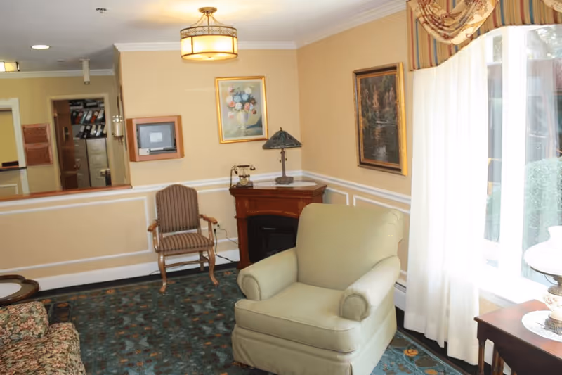 Cozy living room with an upholstered armchair, side table and lamp, framed artwork, and a large window with curtains.