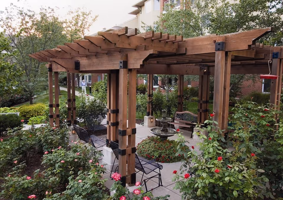 Outdoor seating area with a wooden pergola surrounded by lush greenery and blooming rose bushes. There are metal chairs and a bench around a central water fountain. Trees and a multi-story building are visible in the background.