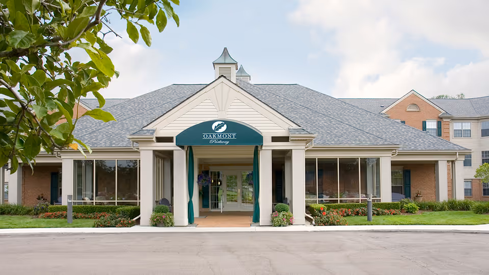 Front exterior view of Oakmont Parkway Senior Community building with a covered entrance, large windows, and landscaped greenery under a partly cloudy sky.