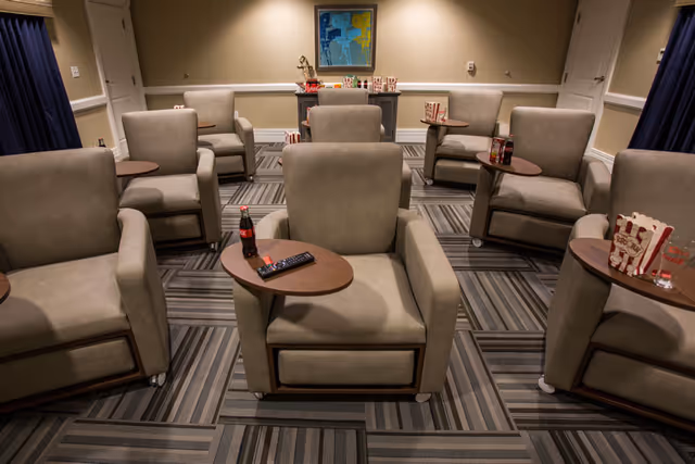 A room with multiple beige armchairs arranged in rows, each with a small attached wooden table. Some tables have snacks like popcorn and drinks. The room has patterned carpet flooring, beige walls, a painting on the far wall, and two closed white doors.