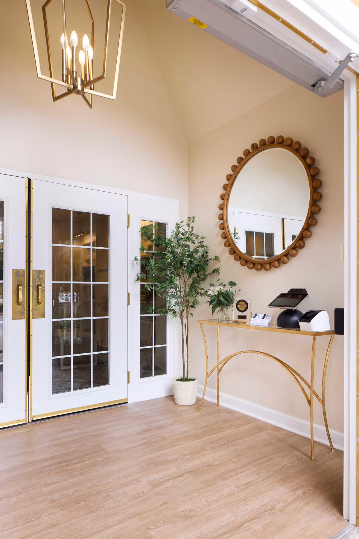 A bright interior corner featuring white double doors with glass panes and gold handles, a tall potted green plant, a gold-framed console table with a small plant and electronic devices on top, and a large round mirror with a decorative wooden frame hanging on the wall above the table. A modern chandelier with exposed bulbs hangs from the ceiling.