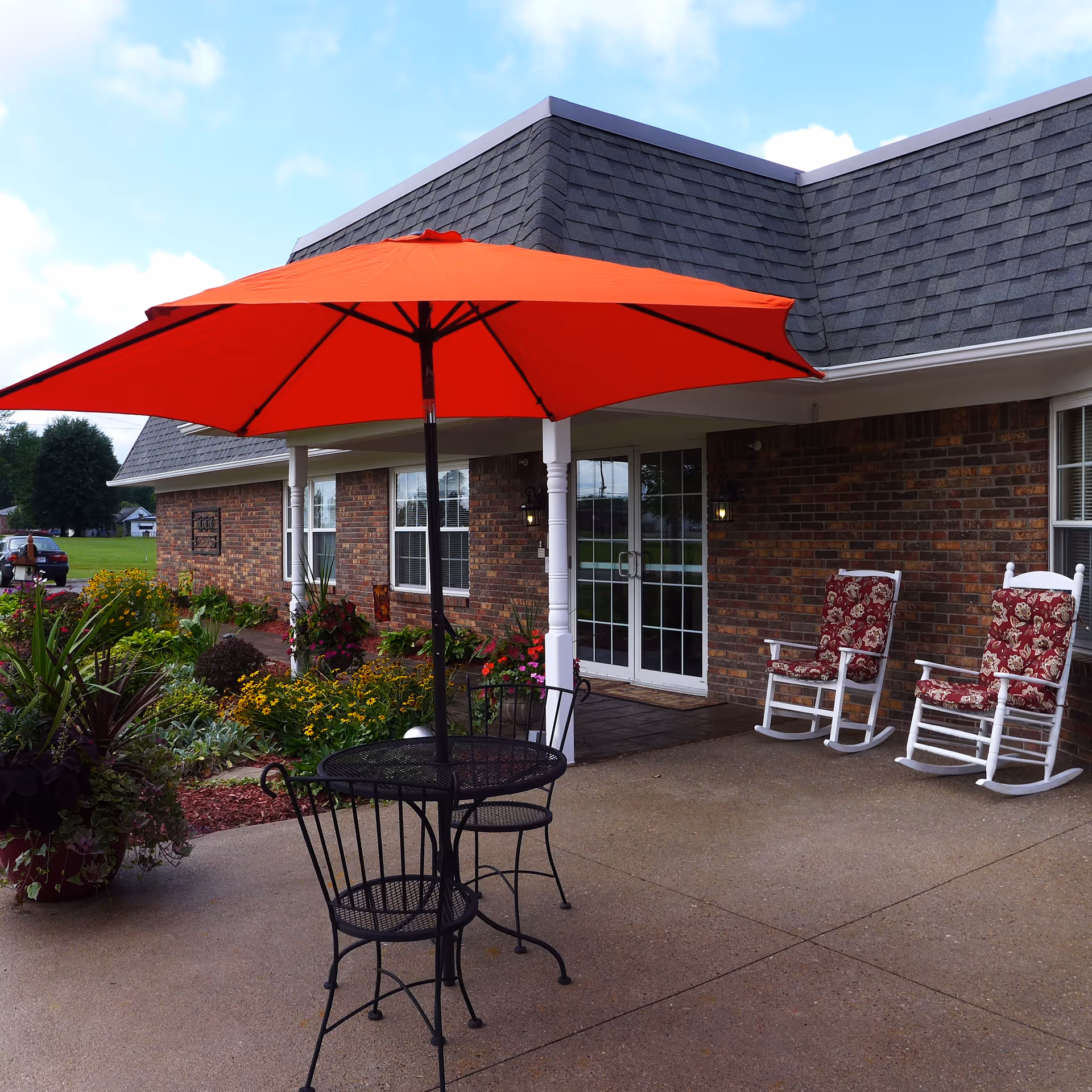 Outdoor patio area at Hickory Creek at Scottsburg featuring a black metal table with two chairs under a large red umbrella, two white rocking chairs with red floral cushions, brick building exterior, and surrounding greenery and flowers.