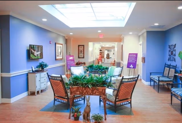 Bright communal seating area in a senior living facility with chairs arranged around planters under a skylight.