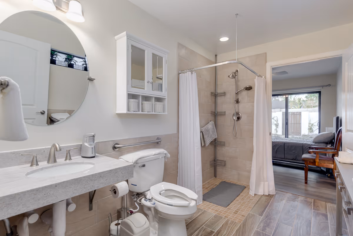 A clean and modern bathroom with a white sink and countertop, a round mirror above the sink, a white cabinet mounted on the wall, a toilet with a towel on top, and a walk-in shower with a white curtain and tiled walls. The bathroom floor has wood-like tiles. Through an open door, a bedroom with a bed, chair, and large window is visible.