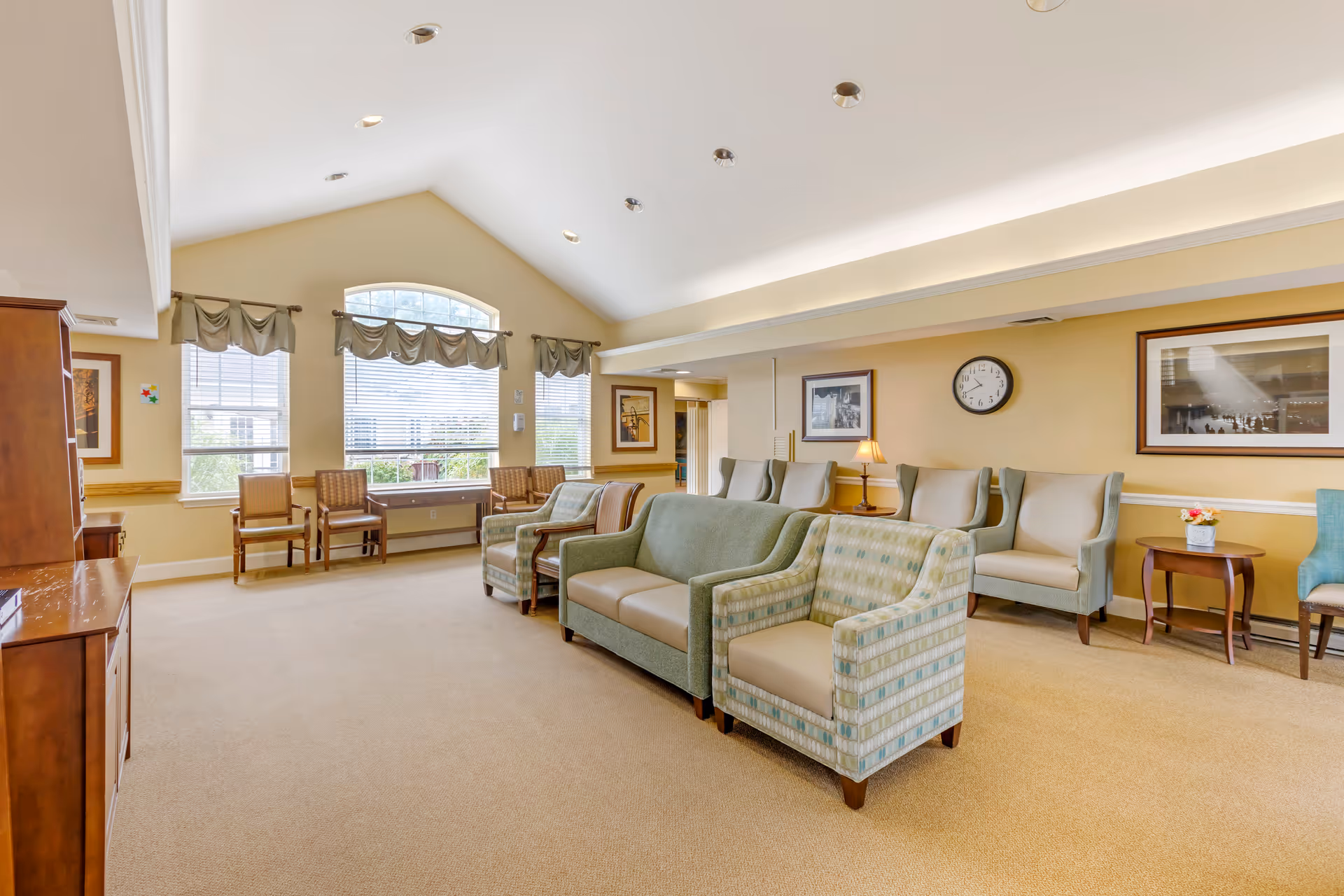 A spacious and well-lit living room area in a senior living facility with beige walls and carpet. The room features multiple upholstered chairs and sofas arranged in rows facing a wooden cabinet on the left. Large windows with valance curtains allow natural light to fill the space. The walls are decorated with framed pictures and a clock. A small side table with a lamp and a flower vase is also visible.